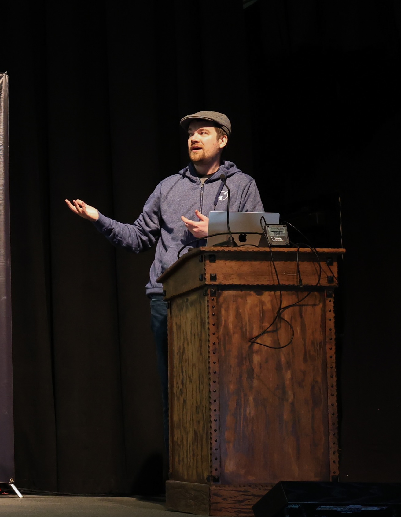 Alex Anderson on stage gesturing at the screen wearing a blue hoodie